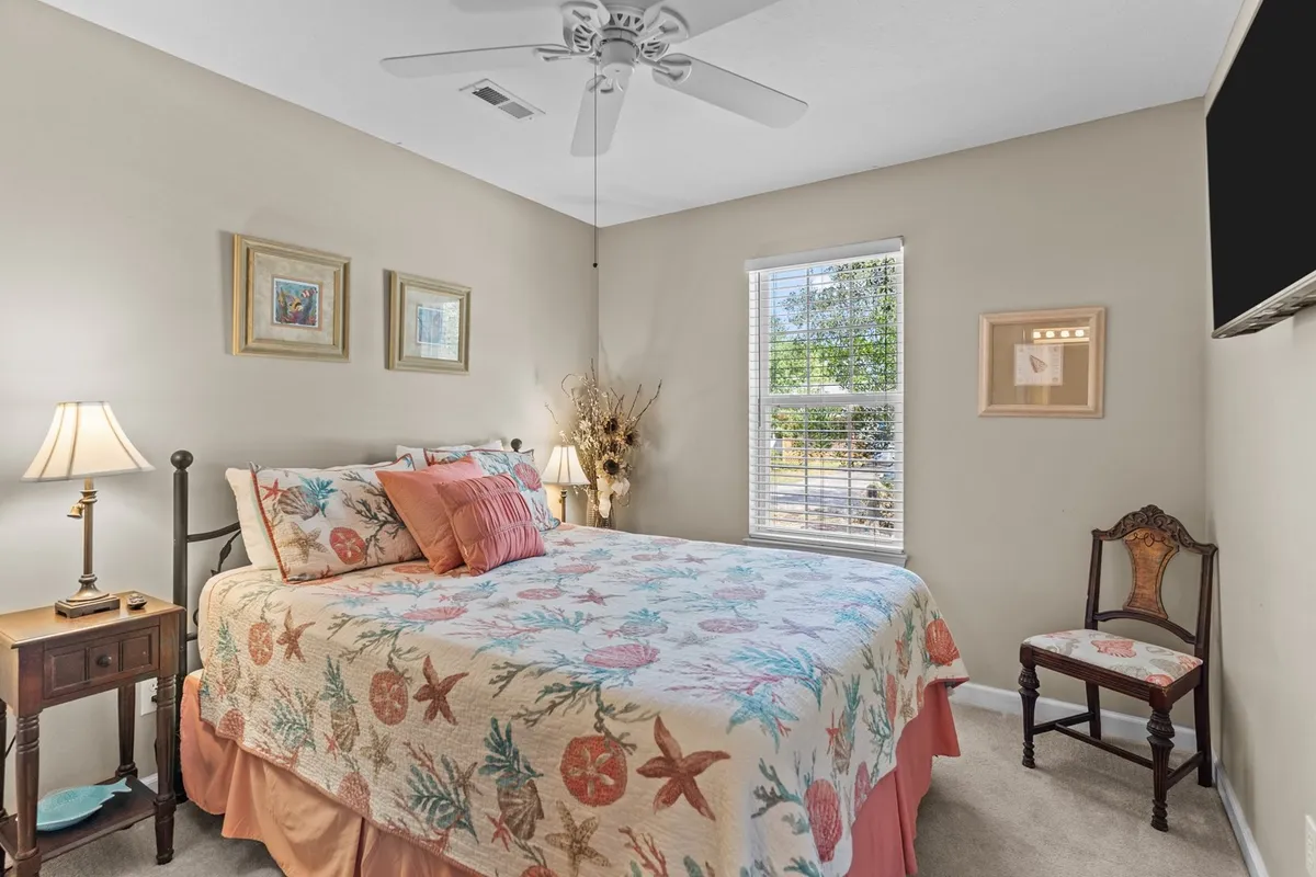 A peaceful bedroom with a bed dressed in a floral quilt, a bedside lamp, a decorative chair, and framed artwork on the wall. The space is filled with soft light, contributing to a tranquil and relaxing ambiance.