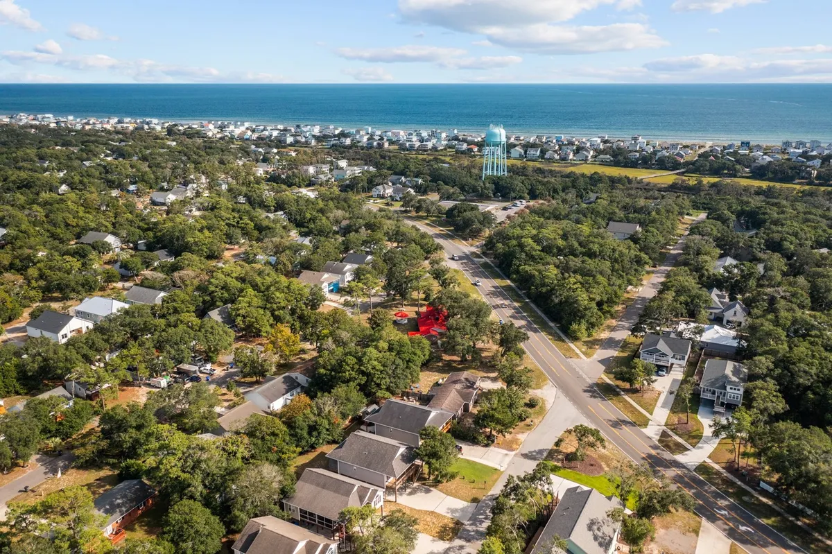 A sweeping view over Oak Island featuring the ocean, water tower, residential homes nestled among a green tree canopy. The image is suffused with relaxed, beach-town energy and highlights the proximity of the ocean to the neighborhood.