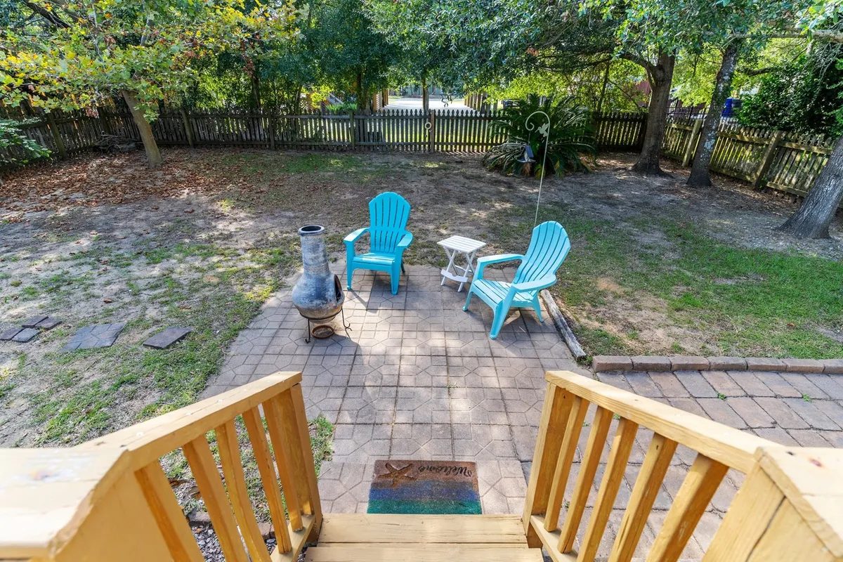 A tranquil patio area featuring two blue Adirondack chairs, a white side table, and an outdoor fire pit, surrounded by greenery. The scene is serene, inviting guests to gather and unwind.