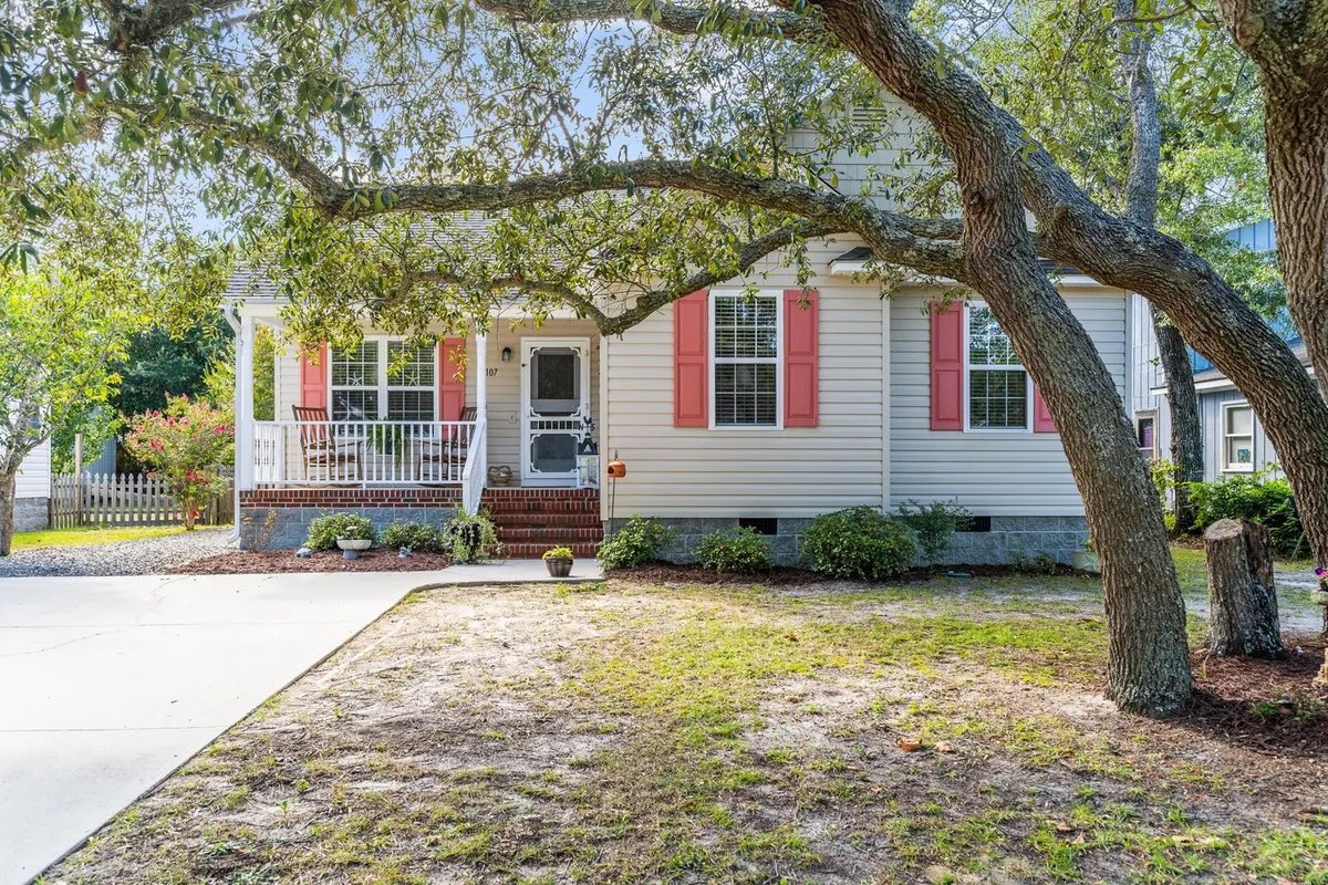 A welcoming exterior view of Shoal Sisters, featuring a front porch with red shutters shaded by a large tree. The scene suggests relaxing porch activities, gardening, and space for family gatherings, evoking a sense of arrival and homely charm.
