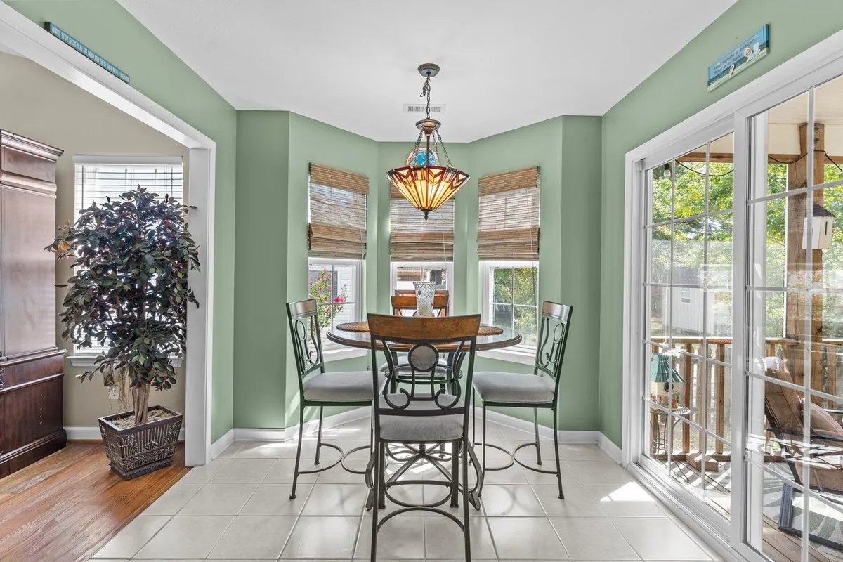 A cheerful dining space highlighted by a decorative chandelier, large windows streaming natural light, and green walls with an indoor plant. The table is set for family meals or casual gatherings, evoking a sense of community and warmth.