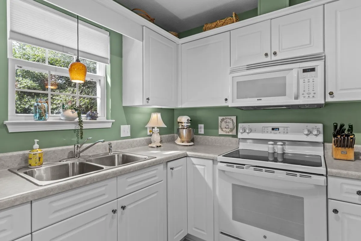 A cozy kitchen scene featuring a kitchen sink and stove, with a window overlooking greenery that lets in natural light. Kitchen accessories and a decorative lamp add a homey touch. The layout showcases the kitchen's functionality and warmth, ideal for family cooking and gatherings.