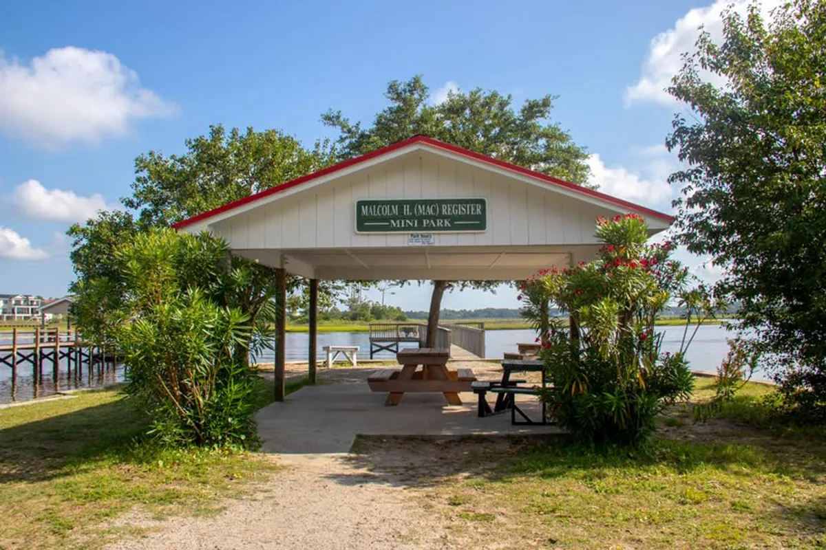 A friendly, communal pavilion with a sign, picnic tables, and views of the water surrounded by greenery. The mood is inviting and social, ideal for family gatherings, picnics, or fishing trips.