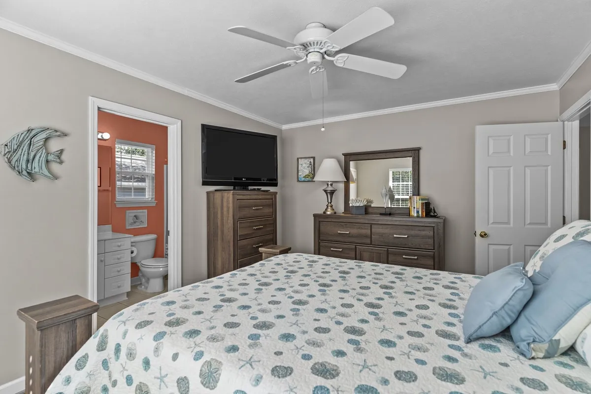 A cozy bedroom featuring a queen-sized bed with a patterned quilt, a dresser with a mirror, a wall-mounted TV, and decorative fish art. The room is softly lit by natural light, creating a welcoming and restful atmosphere.