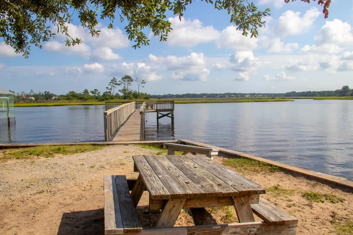 A tranquil dock extends out over calm water, flanked by lush green trees. A wooden picnic table sits nearby, inviting visitors to enjoy meals or simply take in the view. The scene is peaceful, with abundant opportunities for birdwatching, fishing, or watching the sunset.