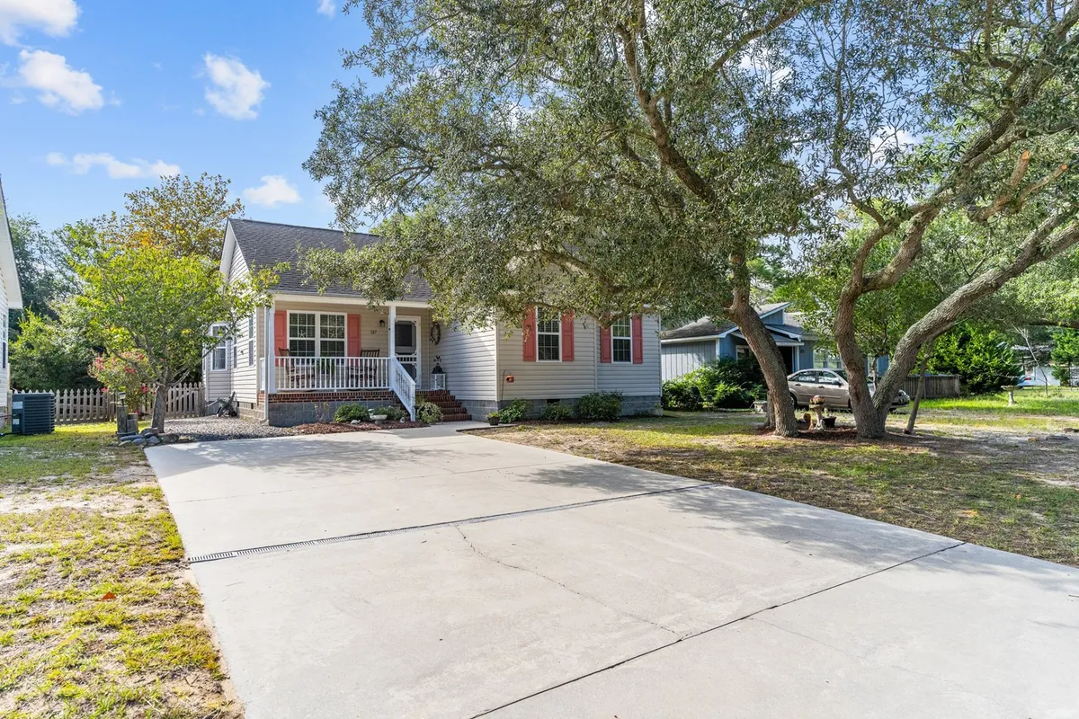 A wide exterior shot emphasizing the front porch, driveway, and large trees surrounding Shoal Sisters. The setting suggests outdoor relaxation, family gatherings, and barbecues, with a warm, welcoming mood.